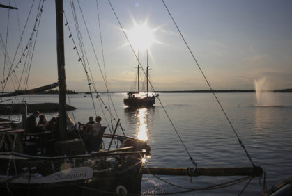 Venesatamassa purjealus rantaan kiinnitettynä ja toinen kauempana purjehtimassa.