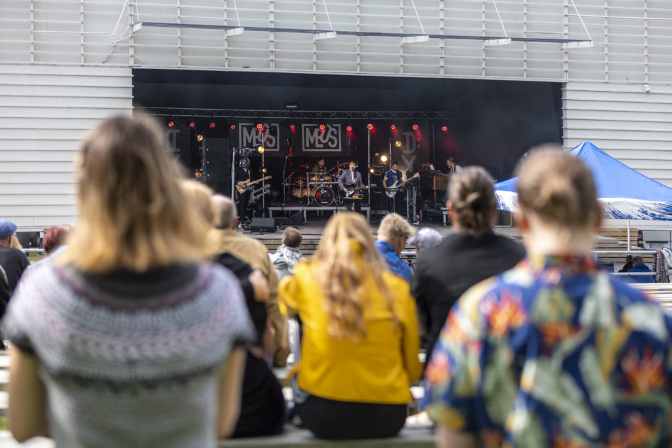 Etualalla istuu selkäpuolelta kuvattuja ihmisiä, taustalla näkyy esiintymislava, jolla soittajia.